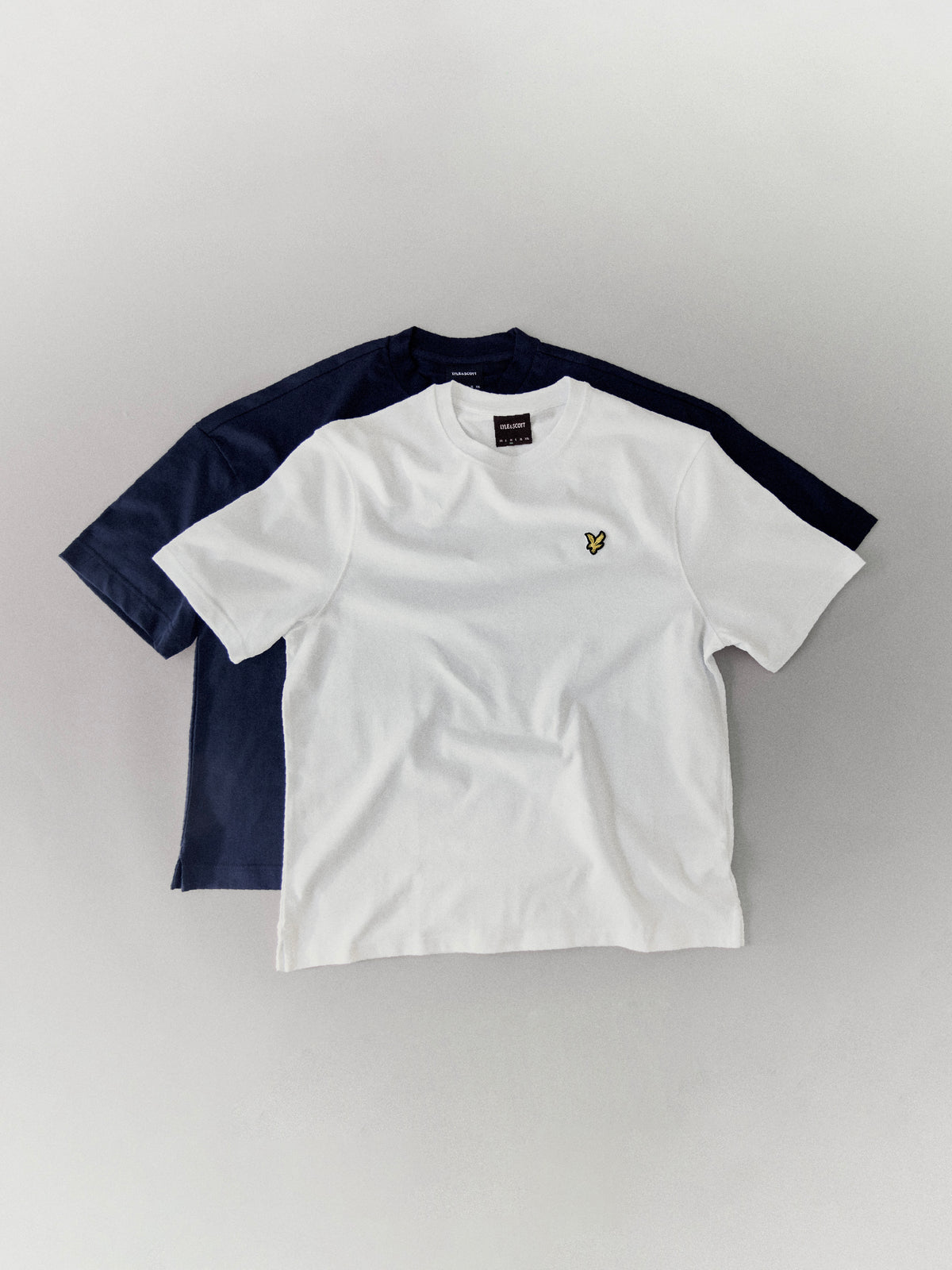 Lyle & Scott - Multipacks - T-shirts, underwear and loungewear. Two short-sleeve crew-neck T-shirts laid flat on a light gray background: a white shirt in front with a small yellow embroidered logo on the chest, and a navy shirt behind it.
