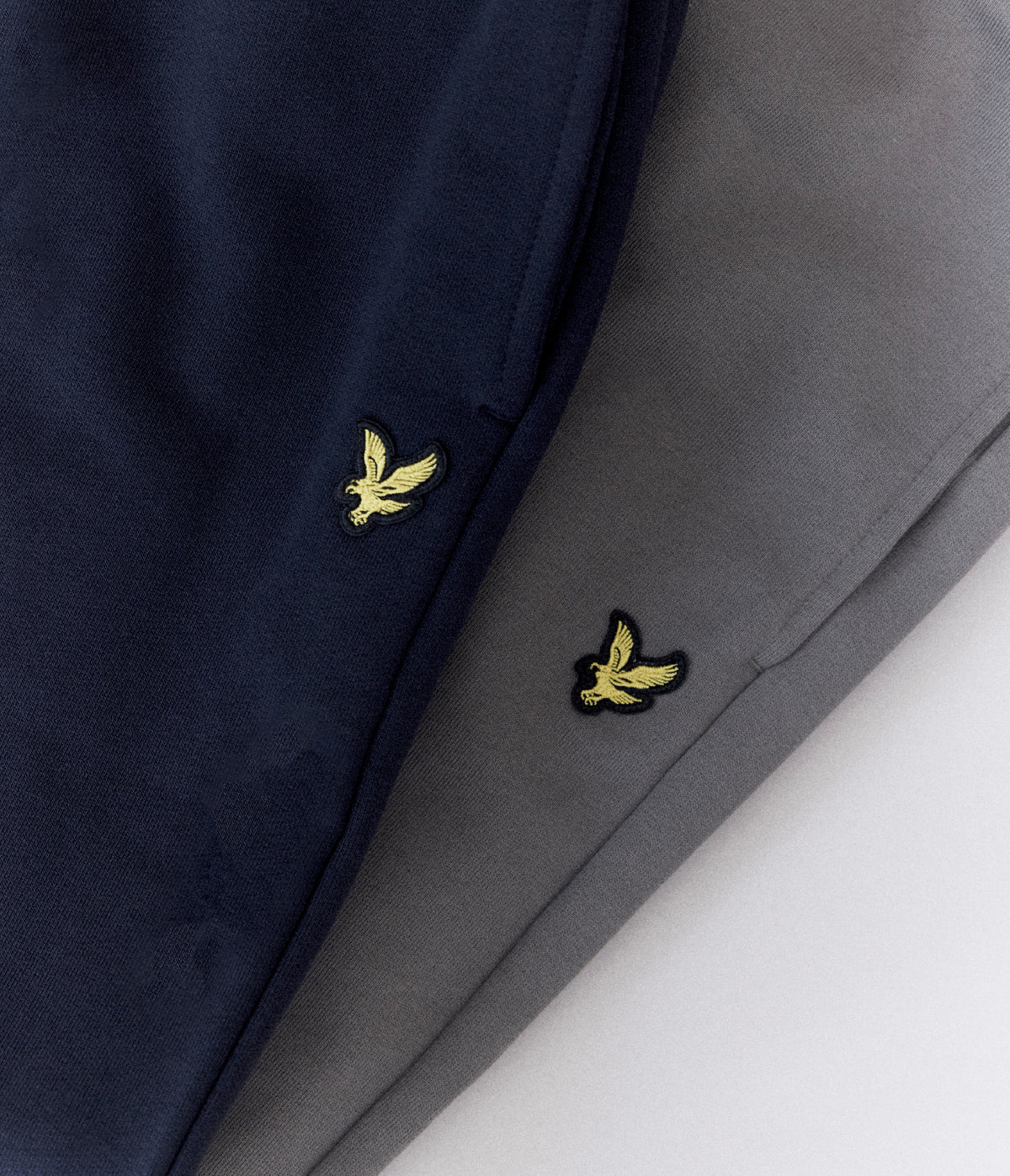 Lyle & Scott - Big & Tall range - 1XL to 5XL. Close-up of folded navy and gray fabric with two small embroidered yellow bird logos, likely pants or trousers, shown on a white background.