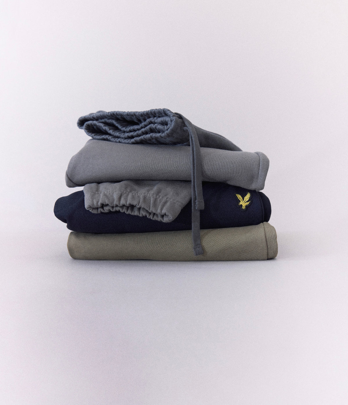 Lyle & Scott - 3 for 2 offer on midlayers - A stack of four folded sweatpants in different colors: slate blue, heather gray, navy blue, and olive green. The navy sweatpants feature a small yellow embroidered Lyle & Scott eagle logo on the side.