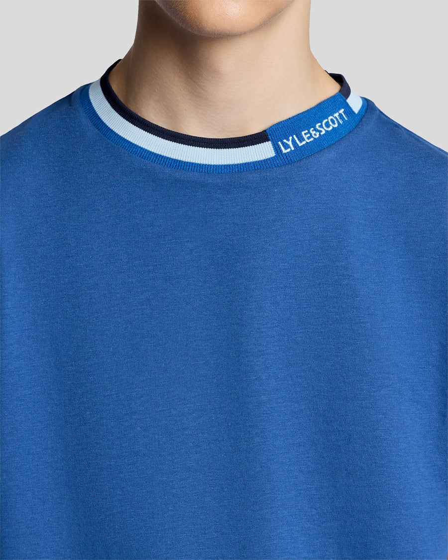 Un homme porte un t-shirt à col rabattu avec logo, couleur bleu barbeau foncé