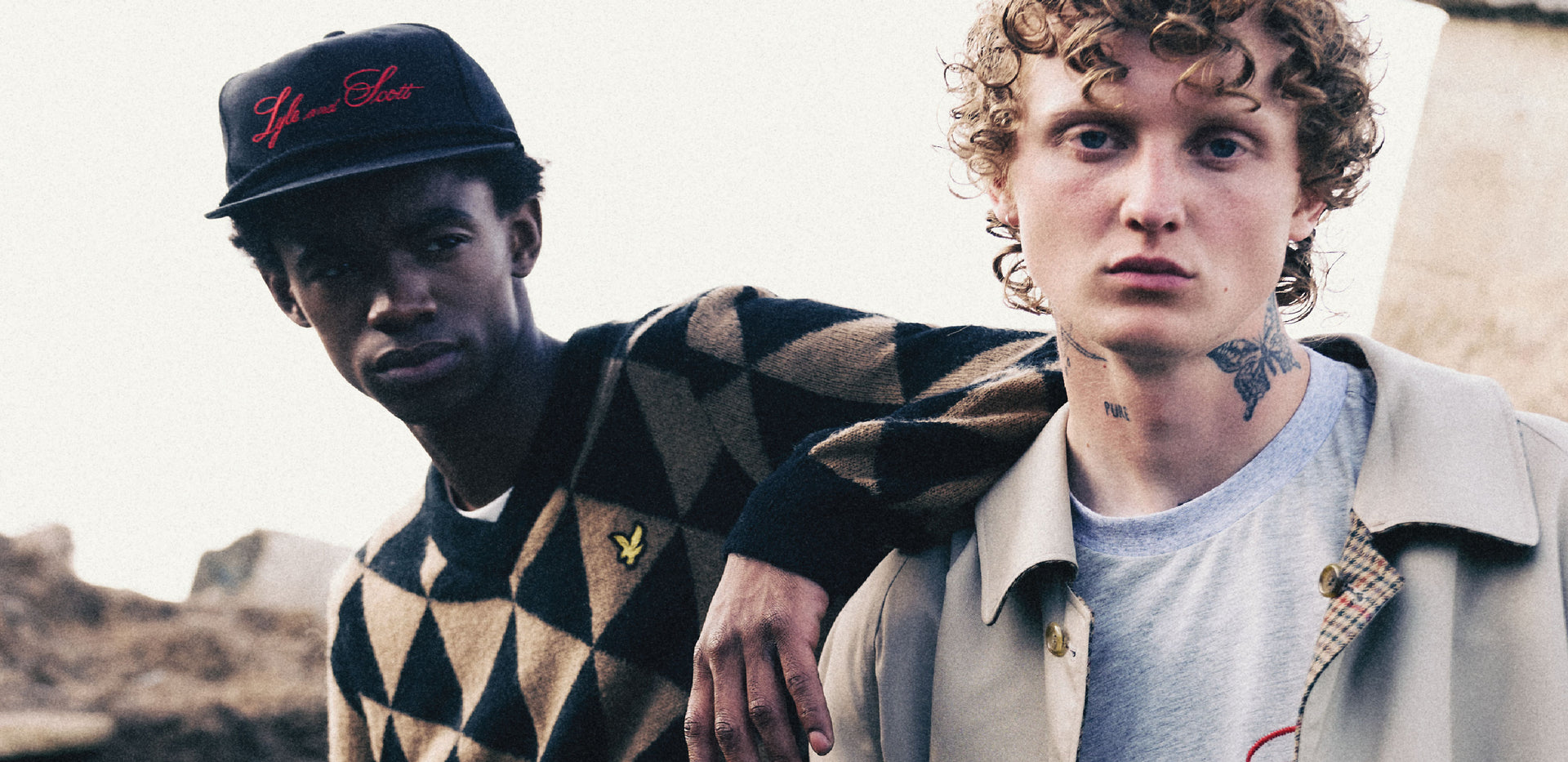 Two men standing close together outdoors, wearing a triangled patterned jumper and hat, or t-shirt.