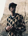 Person wearing a triangle patterned sweater and cap against a textured wall.