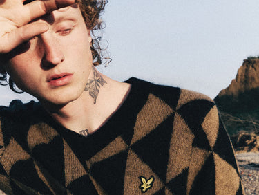 Person wearing a brown and black diamond pattern sweater with an eagle logo, standing outside in sunshine.