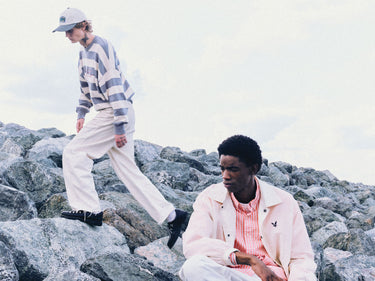 Two people on rocky terrain with a neutral background, wearing a striped jumper and a translucent jacket.