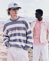 Two people standing on a beach, one wearing a striped sweater with text and a cap, the other in a translucent jacket
